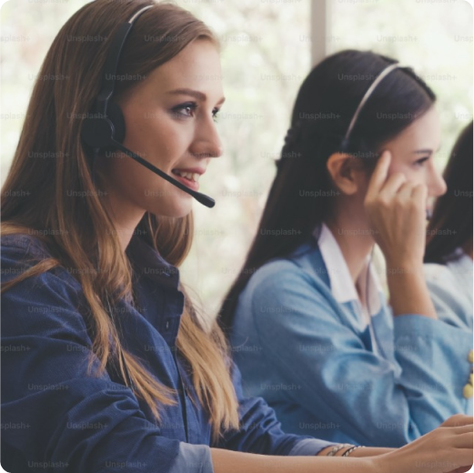 Women with a phone headset on looking at a computer off camera while her and the girl next to her are on the phone.